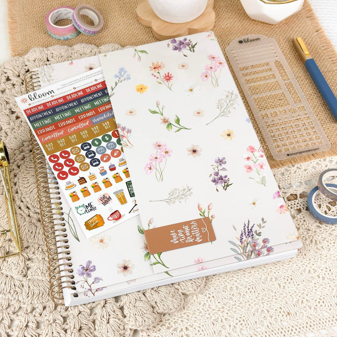 A floral planner sticker sheet and matching sticker pack from Bloom are displayed on a spiral-bound planner, surrounded by stationery props including washi tapes, a ruler, and a pen on a textured beige fabric background.