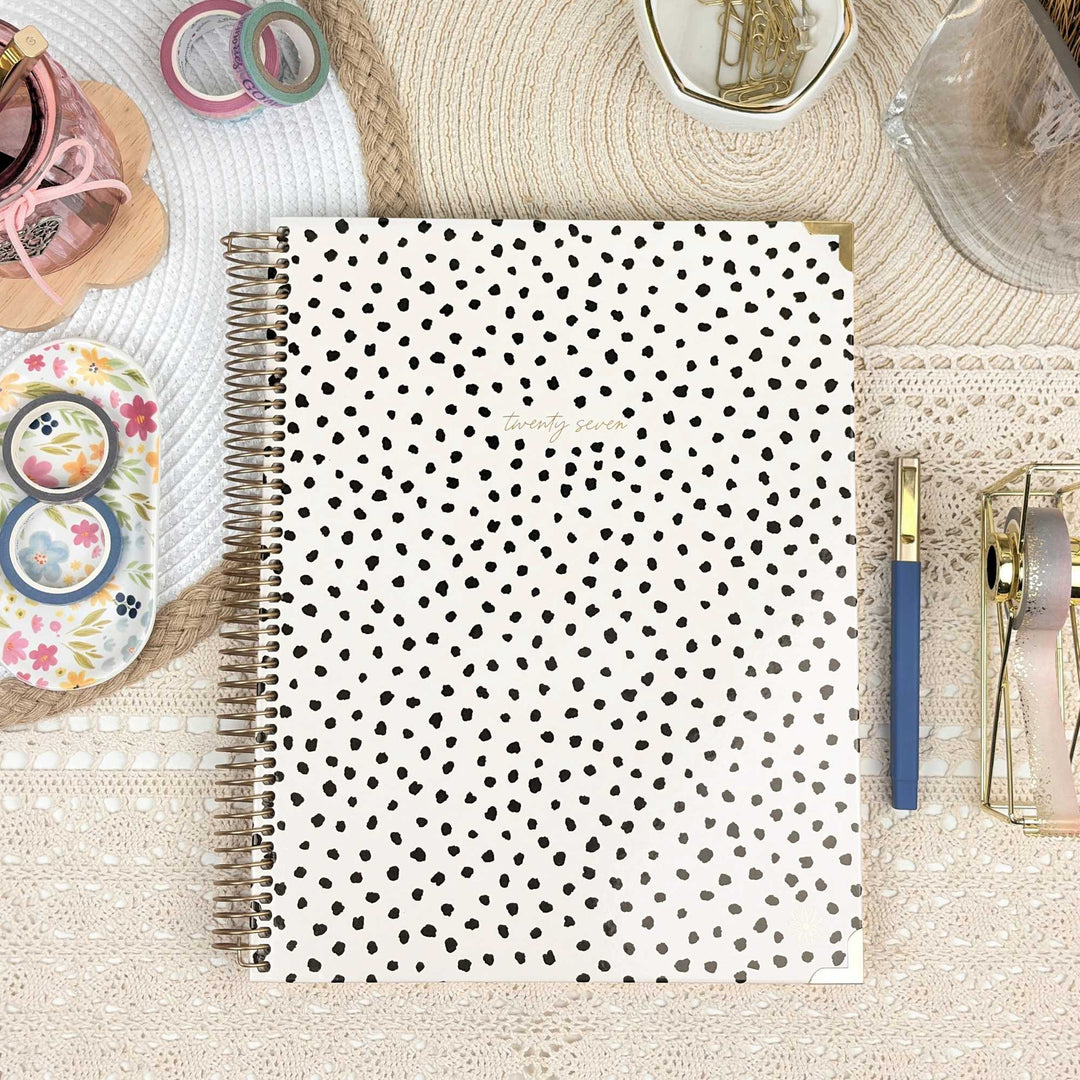 A spiral-bound planner or notebook with a white cover covered in small black hand-drawn dots, styled in a flat lay on a beige lace surface with stationery props around it.