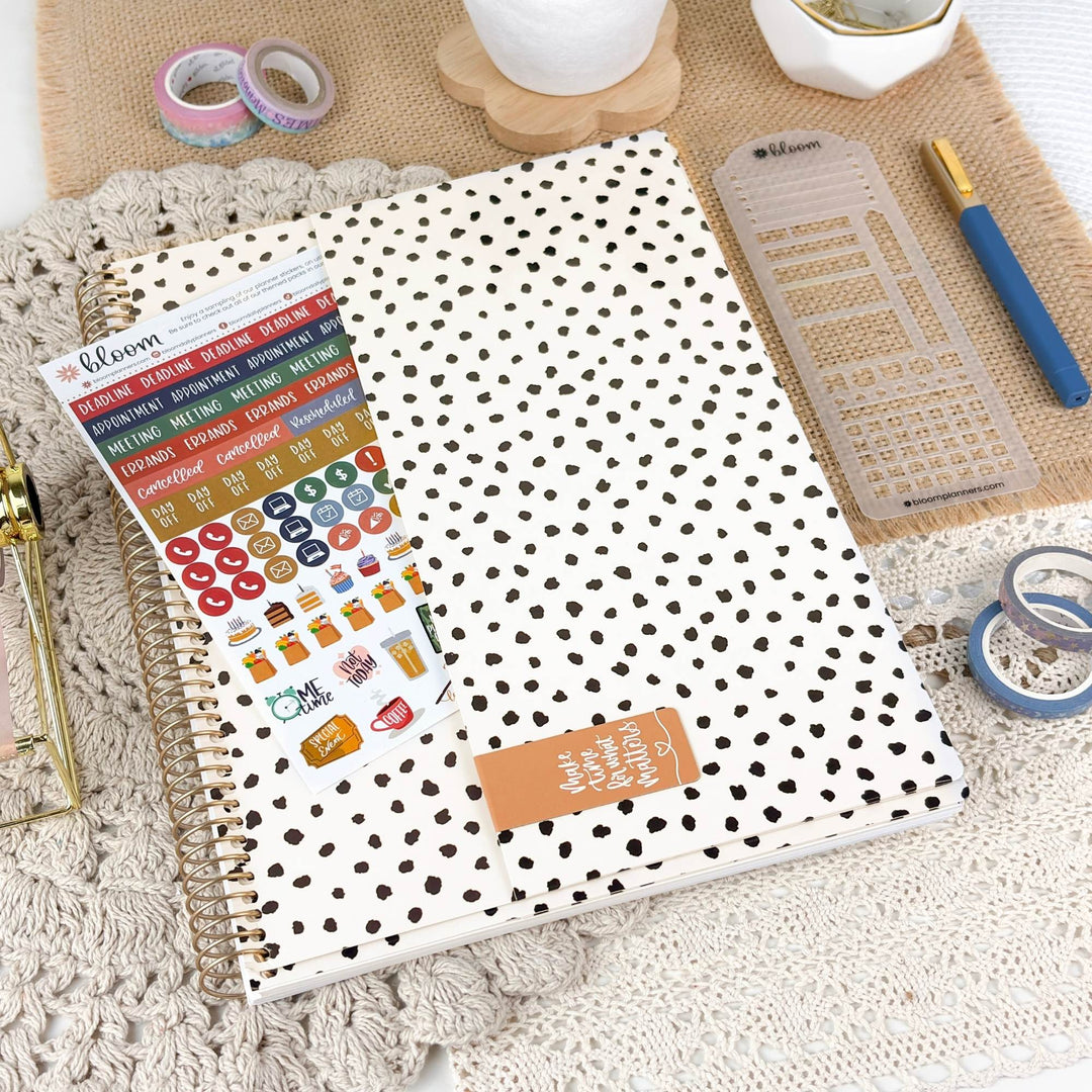 A spiral-bound planner or notebook with a white cover covered in small black polka dots, shown with a sheet of colorful planner stickers. The item is styled in a flat-lay arrangement on a beige textured mat with a blue pen, clear ruler, washi tape, and other stationery accessories.