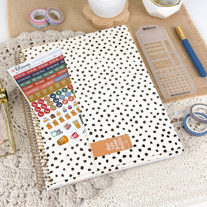 A spiral-bound planner or notebook with a white cover covered in small black polka dots, shown with a sheet of colorful planner stickers. The item is styled in a flat-lay arrangement on a beige textured mat with a blue pen, clear ruler, washi tape, and other stationery accessories.