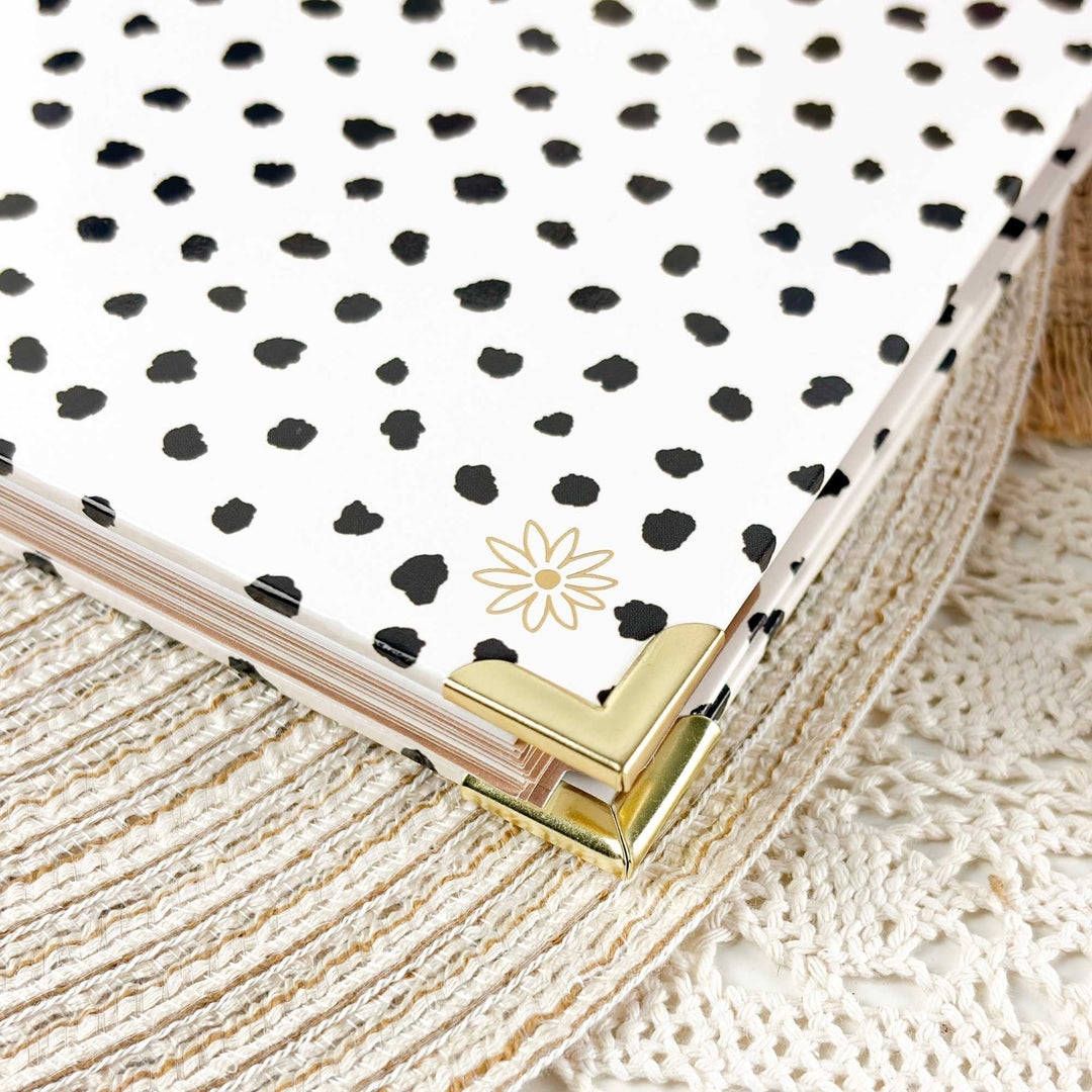 A close-up of a notebook or planner with a white cover covered in irregular black spots, featuring a small gold daisy logo and gold metal corner protectors, resting on a cream lace textile.