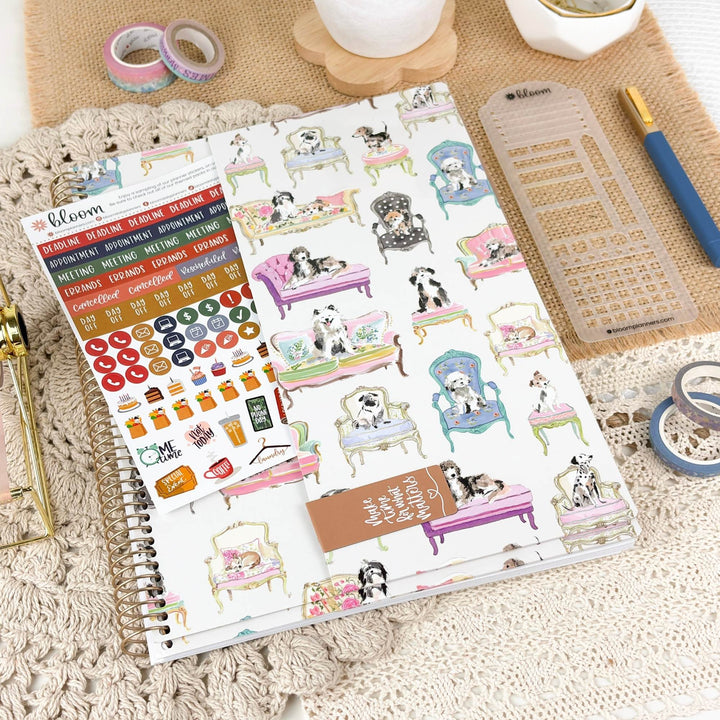 A spiral-bound planner or sticker sheet set displayed on a beige textured surface with stationery props. The main product features illustrated dogs sitting in colorful vintage-style chairs and tables, with a sheet of round and oval stickers and a small brown tag.
