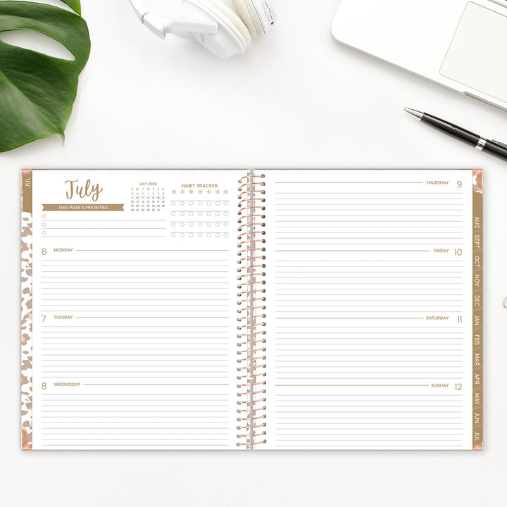 Spiral-bound planner open to a page with July calendar on a white surface with a plant and pen.