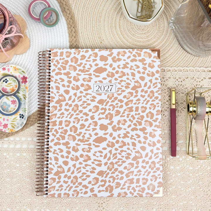 A spiral-bound 2027 planner with a white cover and a light tan leopard print pattern, styled in a flat-lay arrangement on a textured beige surface with decorative stationery props.