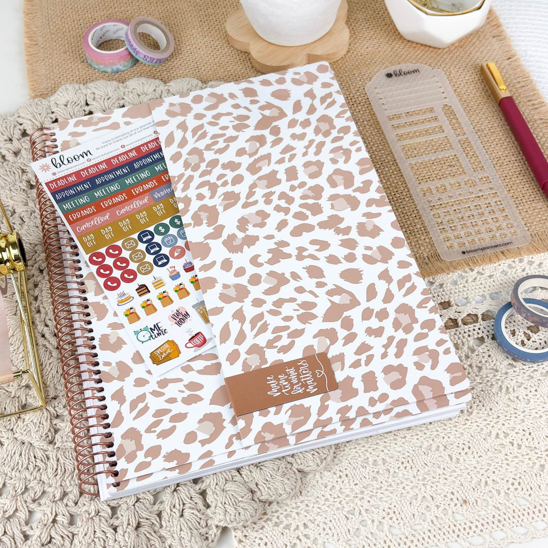 A spiral-bound planner or notebook with a white cover covered in a beige leopard print pattern, shown with a sticker sheet and planner stickers. The notebook is styled on a textured cream lace runner with a burlap mat, washi tape, and other stationery accessories.