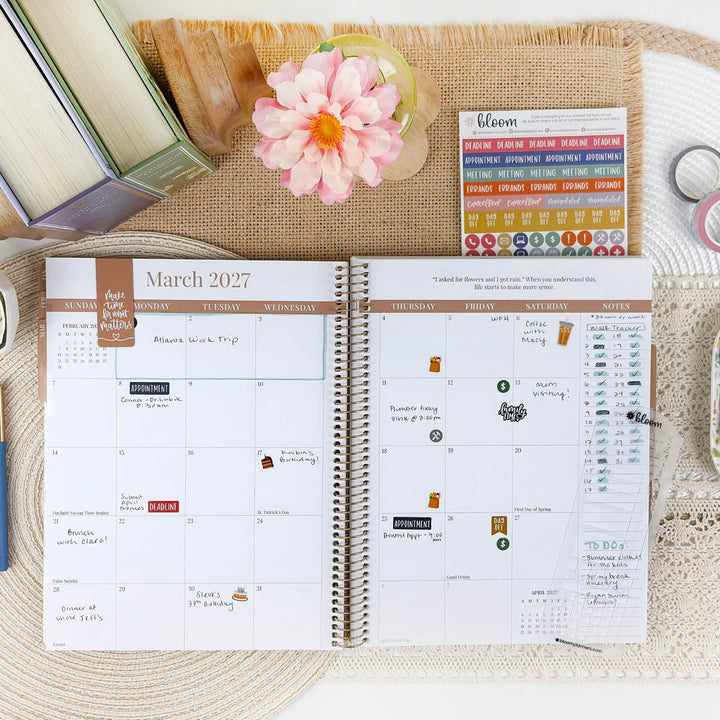 An open spiral planner showing the March 2027 calendar spread, styled on a woven mat with a pink flower, books, and planner stickers around it.