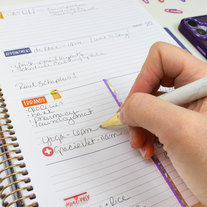Hand writing in a planner with a pencil, surrounded by stationery items.