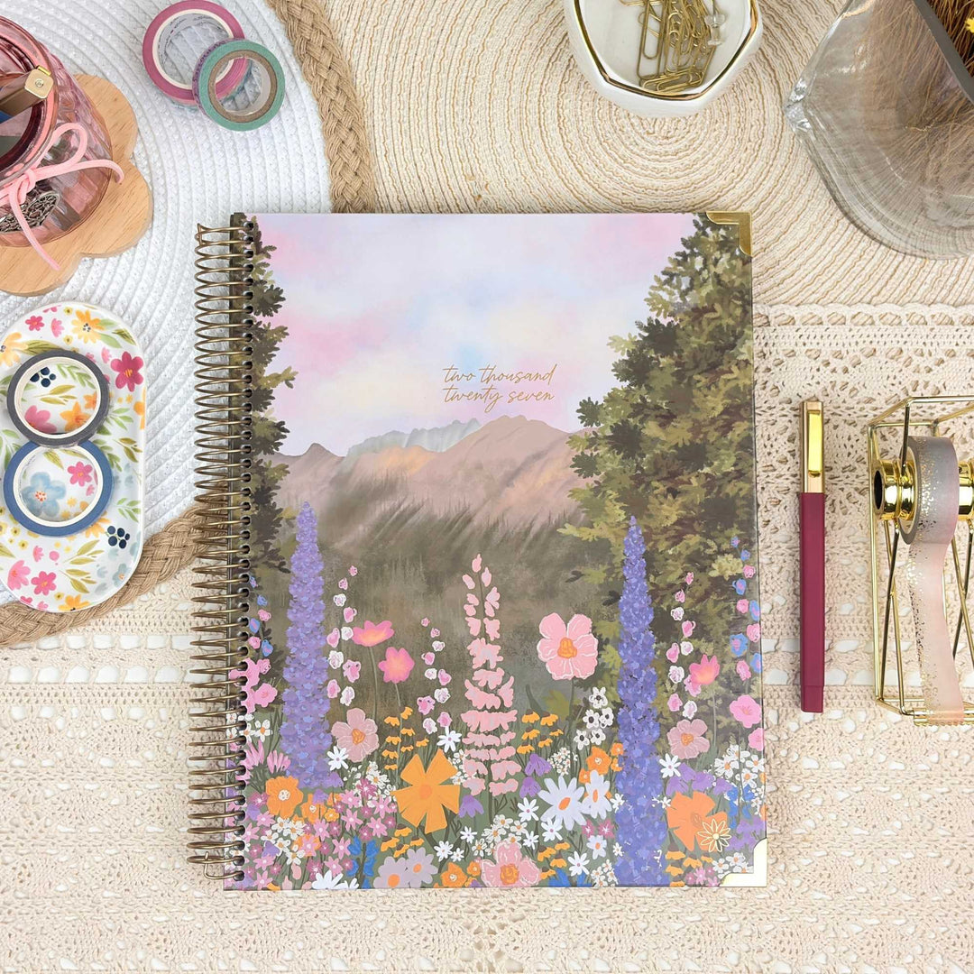 A spiral-bound planner or notebook with a colorful floral mountain landscape cover, styled on a beige lace surface with pens, washi tape, clips, and other stationery around it.