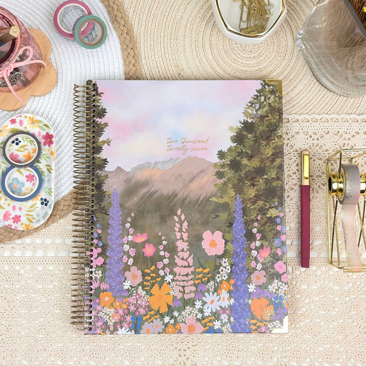 A spiral-bound planner or notebook with a colorful floral mountain landscape cover, styled on a beige lace surface with pens, washi tape, clips, and other stationery around it.