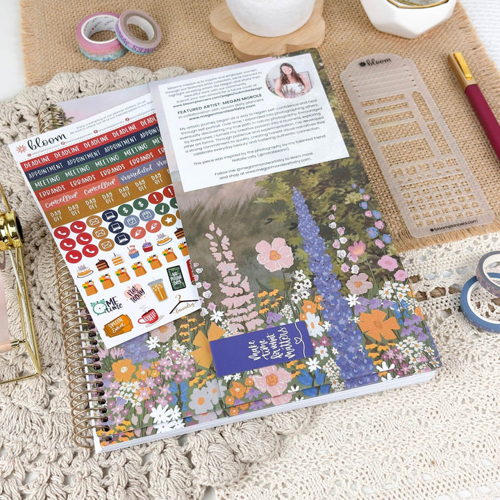 A floral planner or notebook with a colorful wildflower cover lies open on a textured cream lace surface. A sheet of decorative stickers, a clear ruler, a pen, washi tapes, and a small printed card are arranged around it in a styled flat-lay setup.