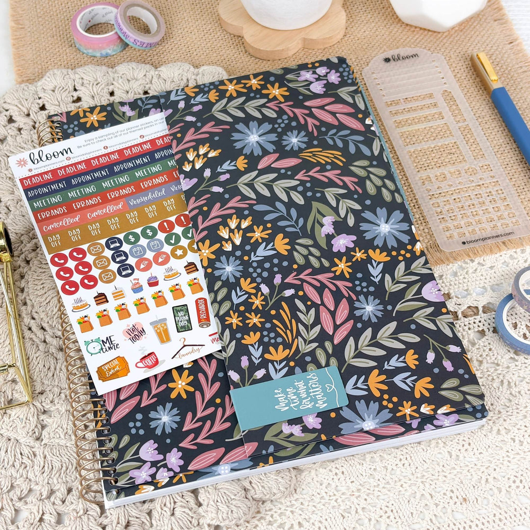 A floral planner or journal cover with a spiral binding, shown in a styled flat-lay with planner stickers, washi tape, a ruler, and a pen on a textured cream surface.