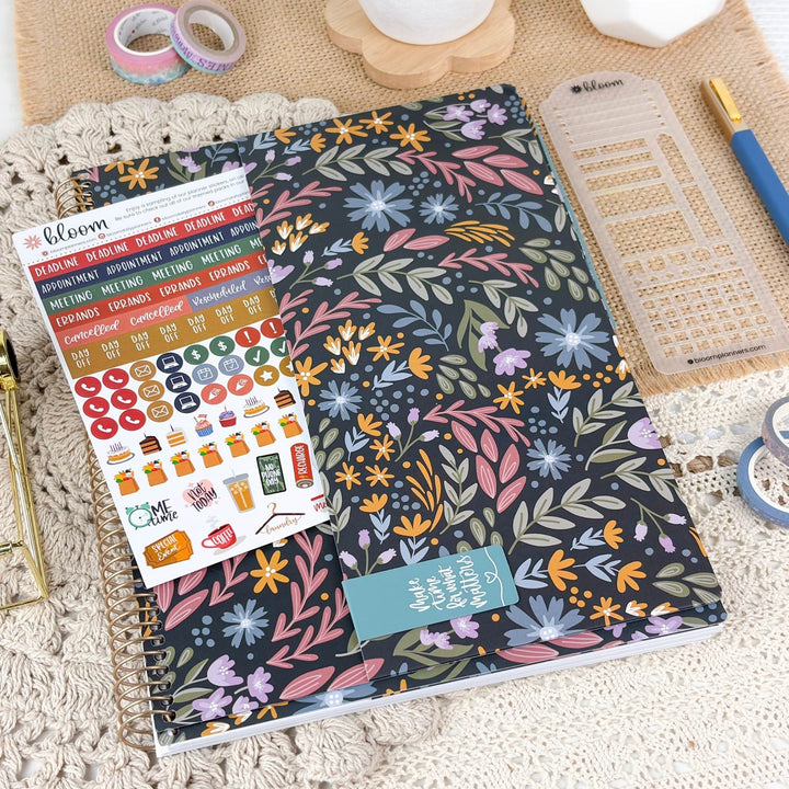A floral planner or journal cover with a spiral binding, shown in a styled flat-lay with planner stickers, washi tape, a ruler, and a pen on a textured cream surface.