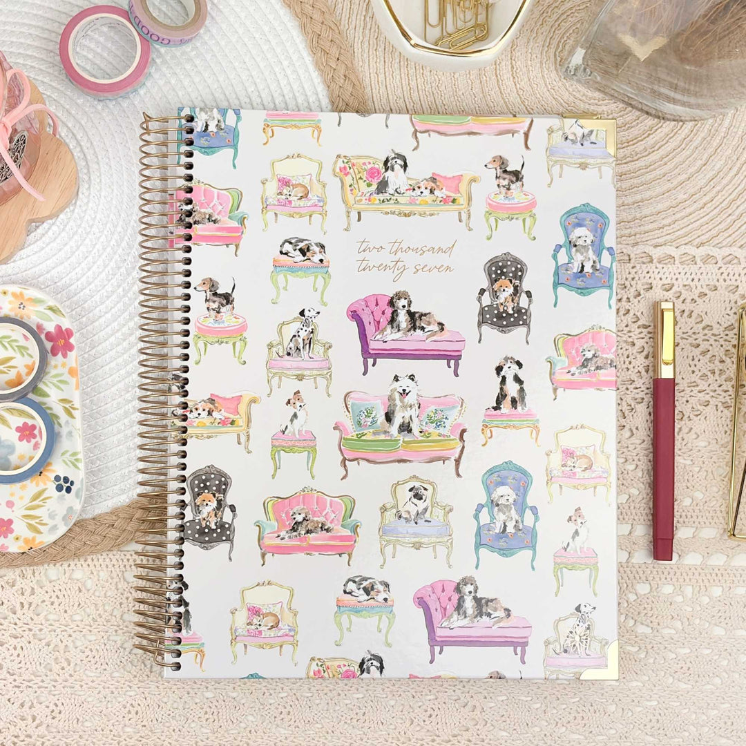 A spiral-bound planner or notebook with a white cover decorated with a colorful pattern of dogs lounging in pink, purple, blue, and green chairs, styled on a lace surface with pens, washi tape, and other stationery around it.
