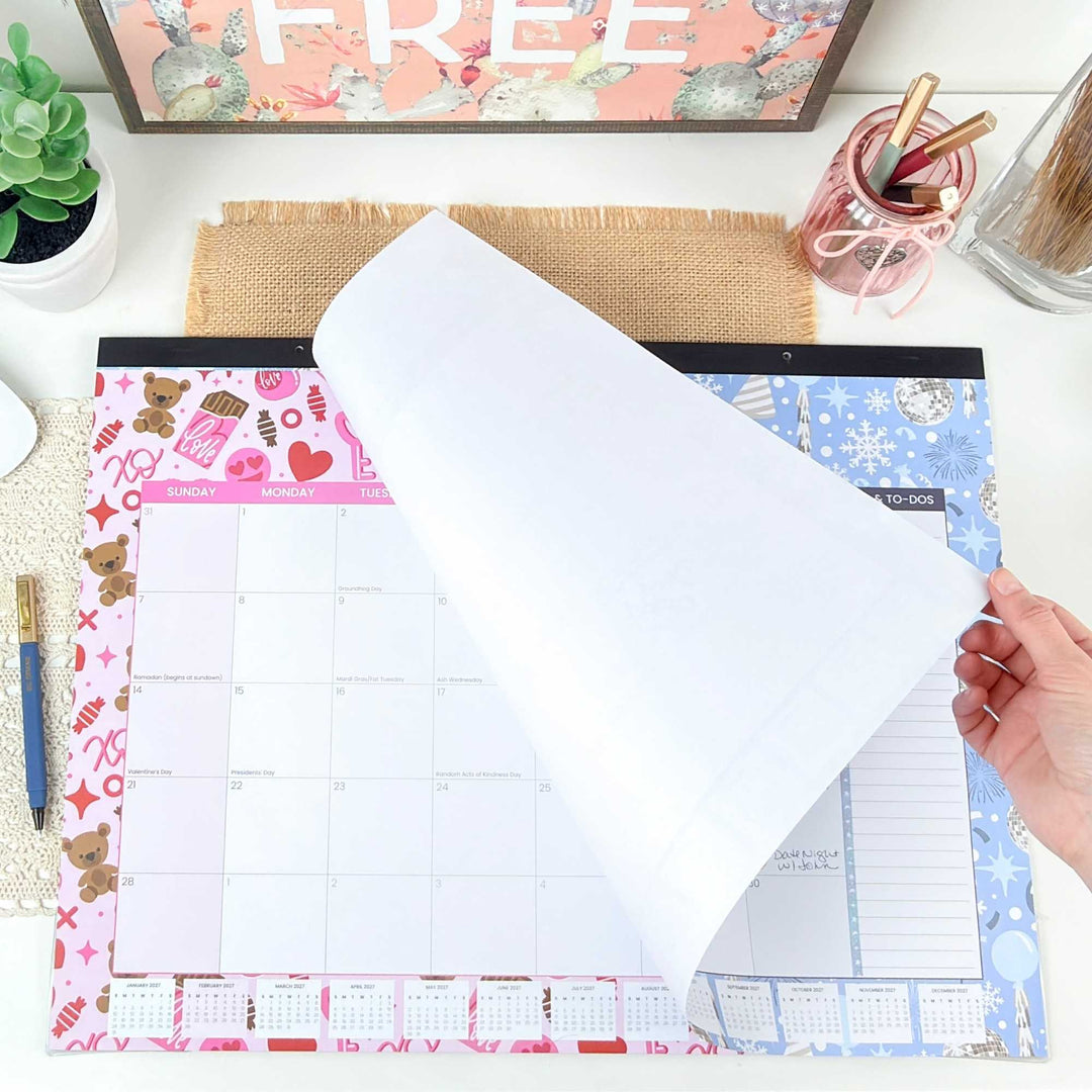 Person holding a transparent sheet over a planner with colorful designs on a desk.