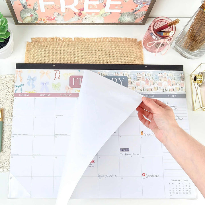Person using a large desk calendar on a desk with office supplies.