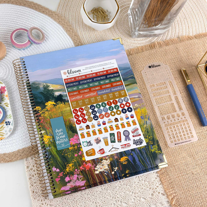 A spiral-bound planner or journal with a colorful floral landscape cover lies on a styled desk. The cover shows a field of yellow, pink, and purple flowers under a blue sky.