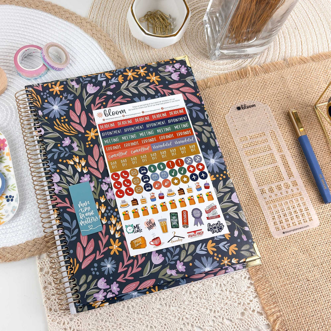 A spiral-bound planner with a dark floral cover lies on a woven mat, surrounded by planner accessories including sticker sheets, washi tapes, a pen, and a toothpick holder. The planner cover features a dense multicolor floral pattern with pink, blue, yellow, green, and purple tones.