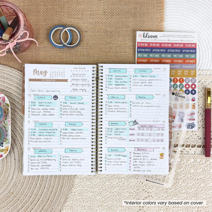 An open spiral-bound planner or journal spread on a woven beige surface, showing weekly planner pages with handwritten notes and colorful headers. Surrounding props include a pink pen, a small jar of pens, two blue rings, a sticker sheet labeled bloom, and a decorative item on the left.