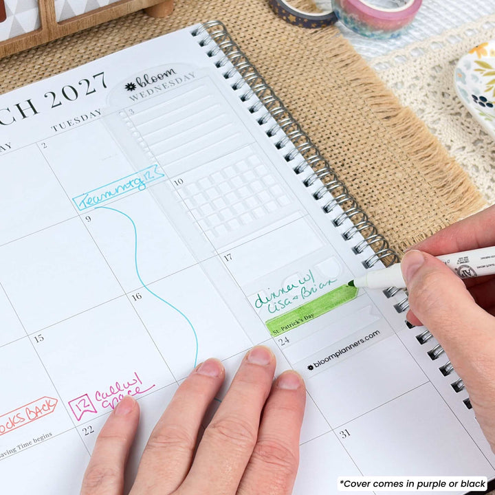 A close-up of a person's hands using a white highlighter to mark a date in a spiral-bound planner. The planner page shows the month of March 2027 with handwritten notes in green, pink, and blue ink.