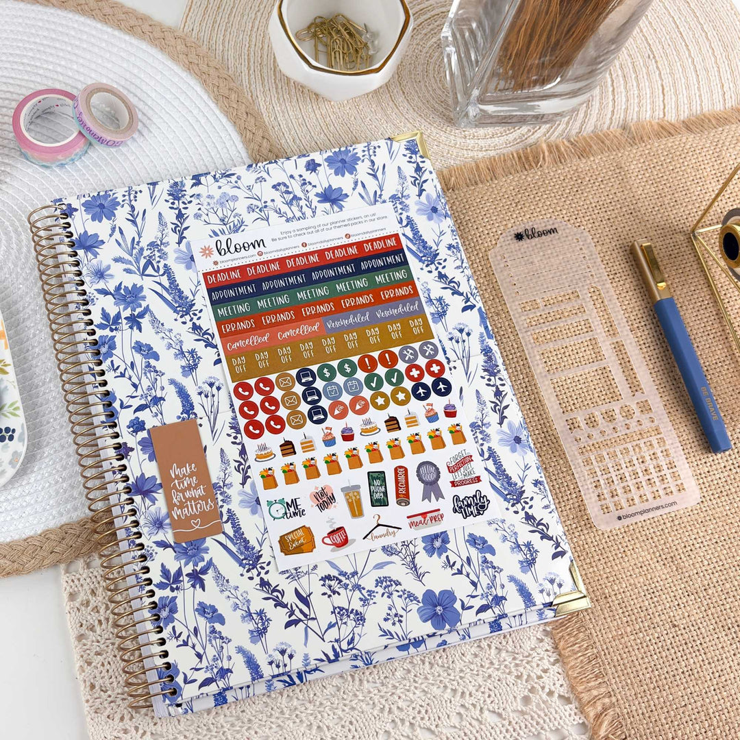 A spiral-bound planner or notebook with a blue floral cover is shown on a styled desk surface, with a sticker sheet attached to the front. The scene includes a pen, a clear plastic grid tool, washi tape, a small dish, and a glass jar in a real tabletop setting, so this is Lifestyle rather than Product/Packaging.