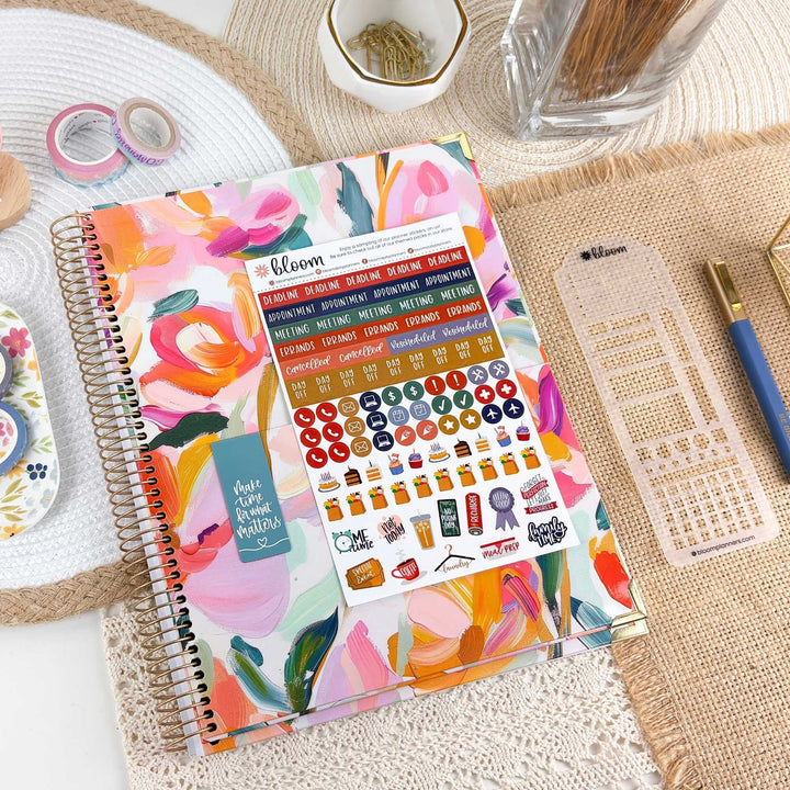 A colorful spiral planner or notebook with a floral watercolor cover and a sheet of planner stickers placed on top, styled on a woven mat with pens, washi tape, and other stationery around it.