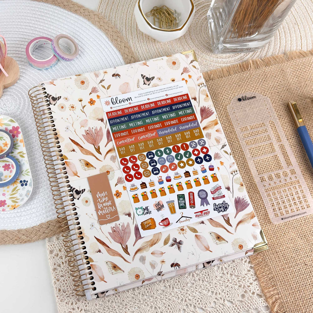 A floral planner or notebook with a spiral binding and a colorful sticker sheet attached to the cover, styled on a woven mat with pens, washi tape, and other stationery around it.