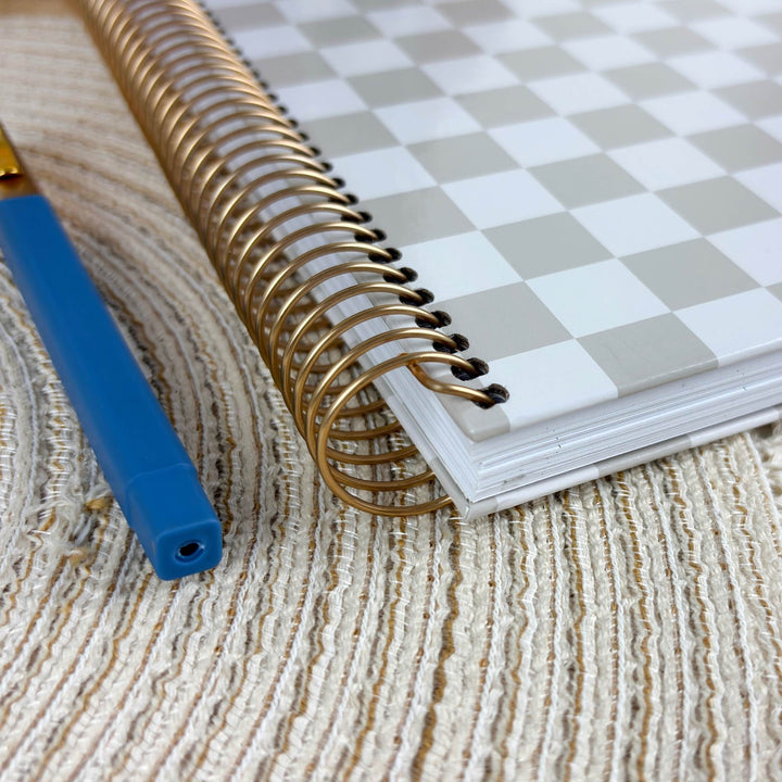 A spiral-bound notebook with a white and light gray checkerboard cover lies on a textured beige woven surface, next to a blue pen with a gold tip.