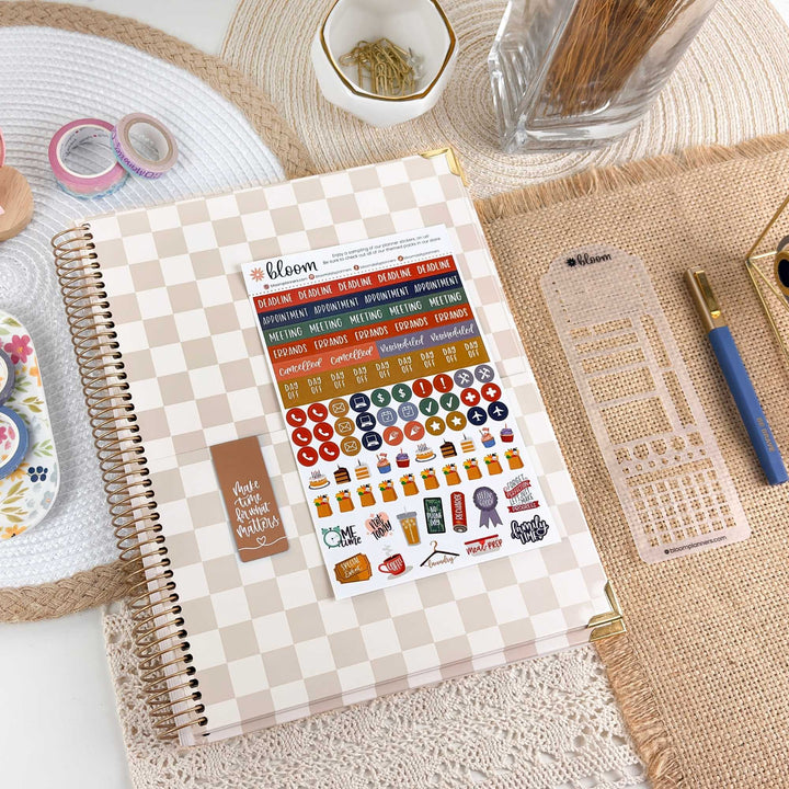 A spiral-bound planner or notebook with a beige-and-white checkered cover lies open on a burlap mat, displaying sticker sheets and a planner insert. Surrounding props include a pen, paper clips, washi tape, and a small dish of flowers in a styled desk setup.