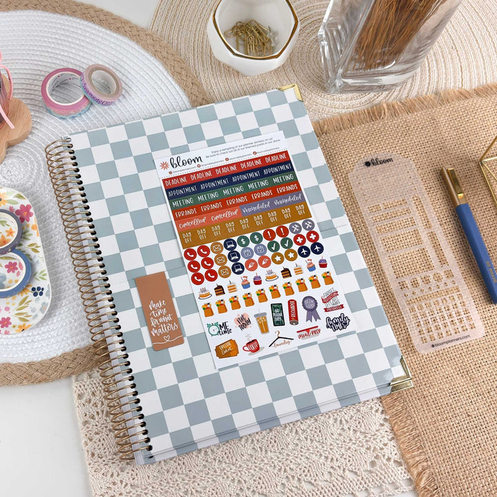A spiral-bound planner or notebook with a blue-and-white checkered cover lies on a woven mat, surrounded by stationery items including washi tapes, a pencil, a clear plastic calendar insert, and small decorative accessories. The cover shows the brand name Bloom and a calendar-themed sticker sheet with colorful icons and text.