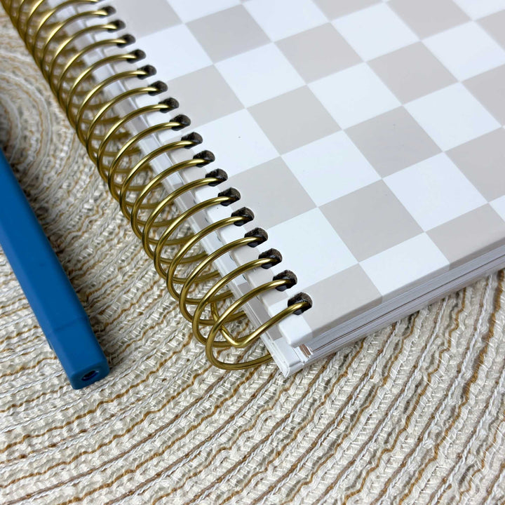 A spiral-bound notebook with a white and light gray checkerboard cover and shiny gold wire binding, placed on a textured beige woven surface next to a blue pen.