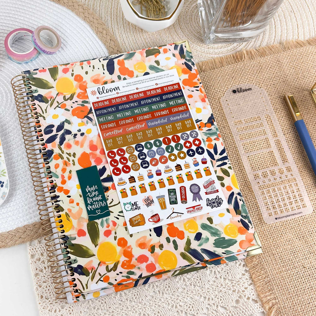 A spiral-bound planner or notebook with a colorful floral watercolor cover is styled on a burlap mat with planner stickers, washi tape, a pen, and other stationery around it.