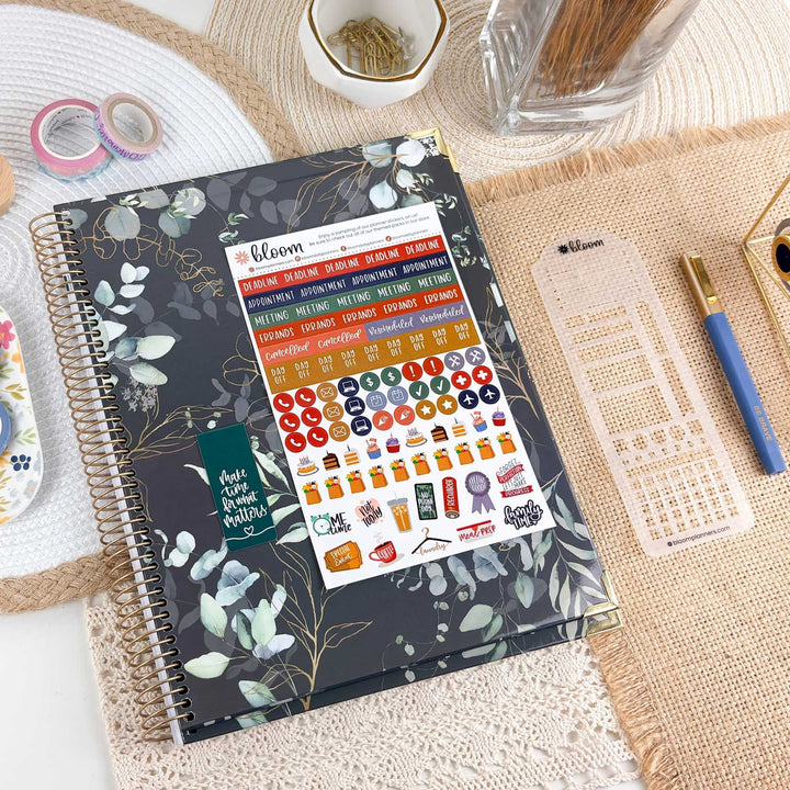 A spiral-bound planner or journal with a dark floral cover lies on a woven mat, surrounded by desk accessories including washi tape, a glass jar, a pen, a clear plastic grid insert, and a small decorative item. The cover shows the bloom logo and a sheet of colorful planner stickers attached to the front.