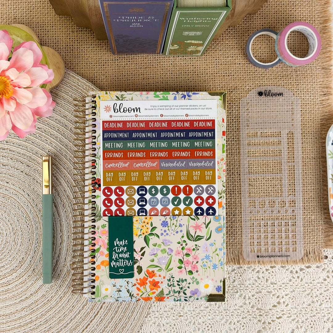 A floral spiral-bound planner labeled 'Bloom' is laid flat on a burlap surface with decorative props including a pink flower, a green pen, washi tape, and a clear plastic sheet. The planner cover shows colorful stickers, a calendar strip, and a green note card that reads 'Your life matters'.