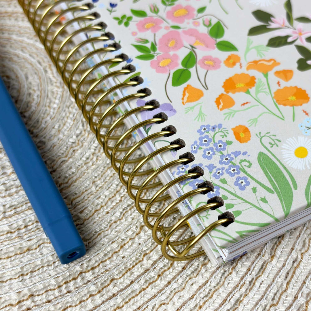 A close-up of a spiral-bound notebook with a white cover decorated with colorful floral illustrations, placed on a woven beige surface next to a blue pen.