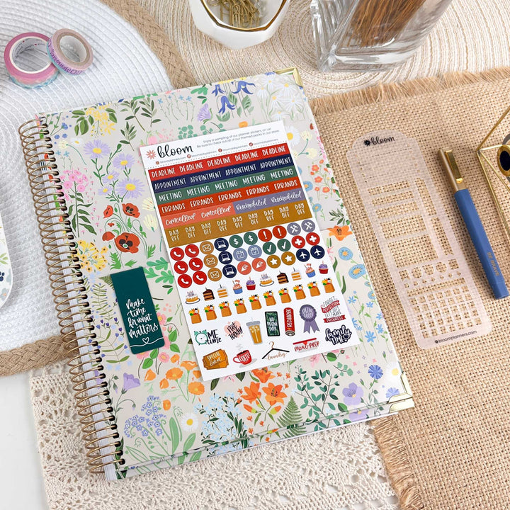 A floral spiral-bound planner or notebook cover with a sheet of colorful stickers placed on top, styled on a woven mat with washi tape, a glass jar, a pen, and a ruler nearby.