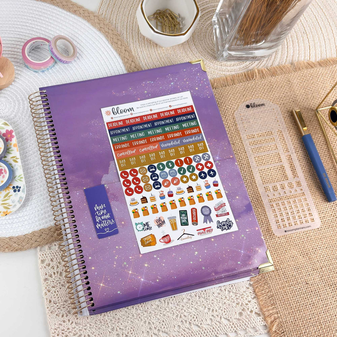 A purple spiral-bound planner or notebook with a sheet of colorful planner stickers applied to the cover, styled on a woven mat with pens, washi tape, and other desk accessories around it.