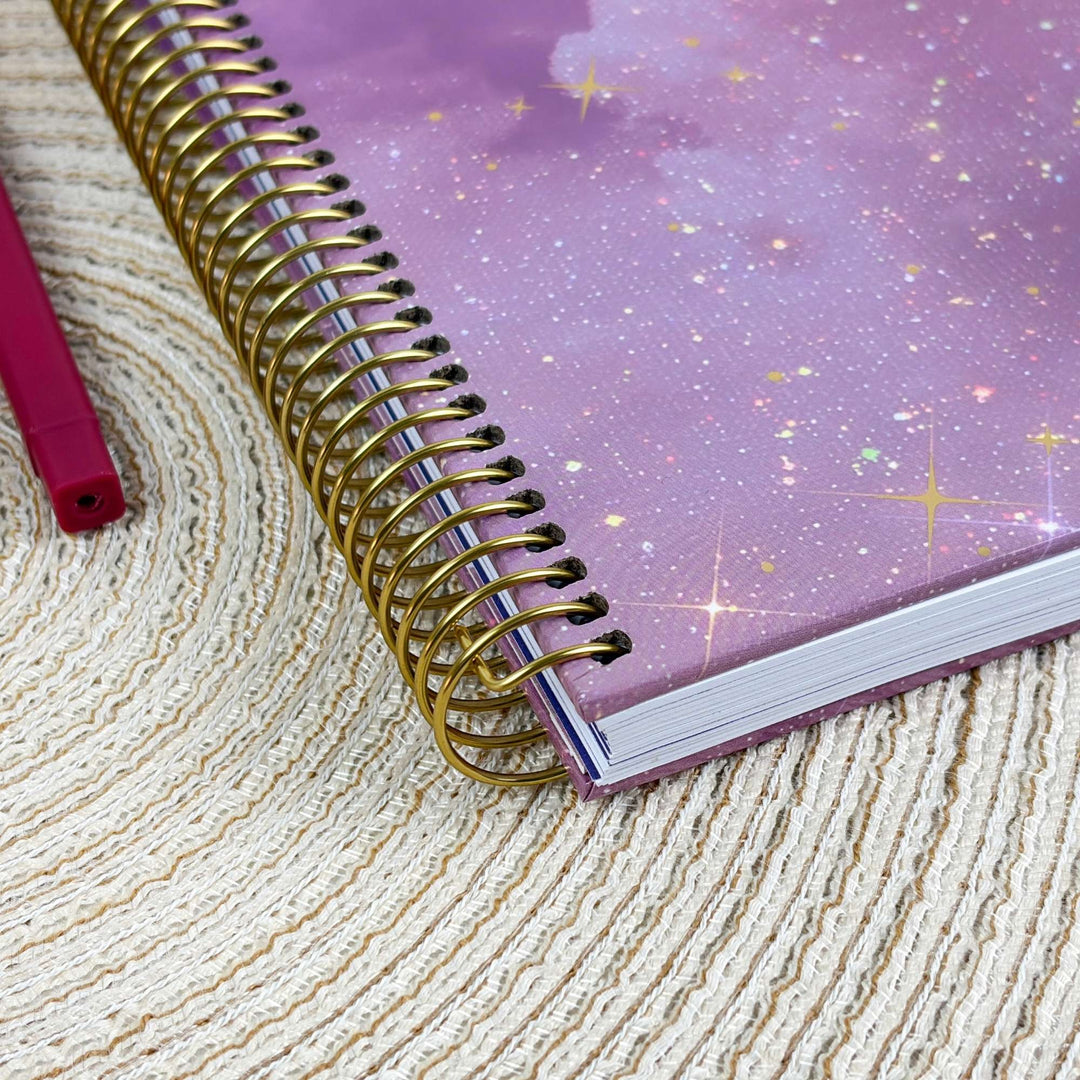 A spiral-bound planner with a lavender-purple galaxy cover and a gold wire coil, placed on a textured beige woven mat next to a pink pen.