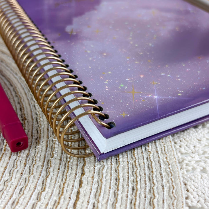 A spiral-bound notebook with a purple galaxy-patterned cover and gold wire binding, placed on a textured cream woven blanket next to a pink pen.