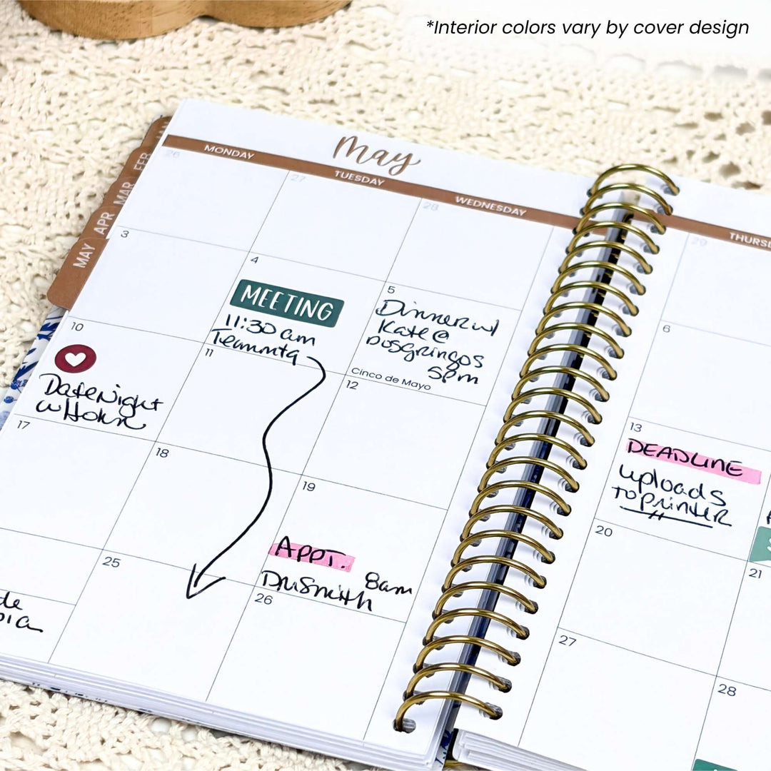 An open spiral-bound planner showing a May calendar page with handwritten notes and a gold wire coil, photographed on a textured cream fabric background. Small text at the top says 'Interior colors vary by cover design.