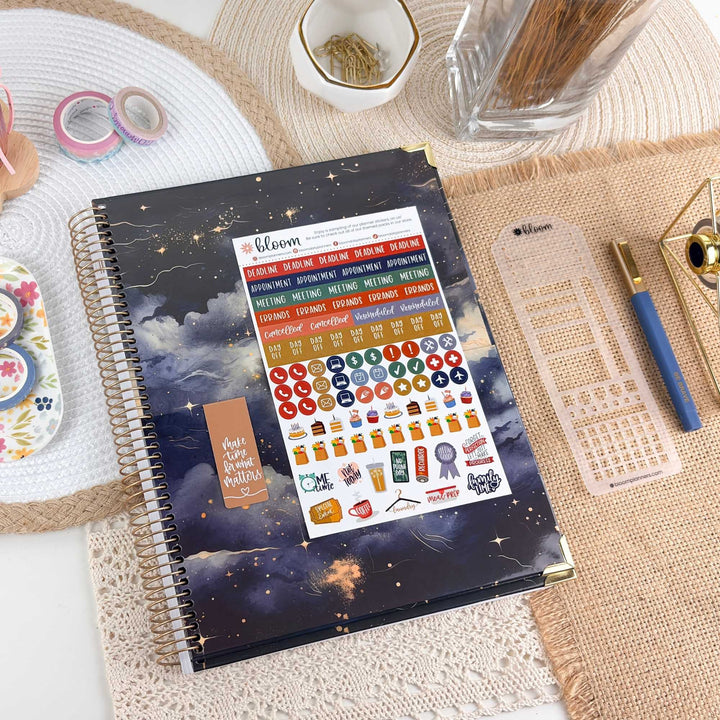 A spiral-bound planner or notebook with a dark blue and black starry sky cover, shown open to a sticker sheet labeled 'Bloom' on a styled desk surface with pens, washi tape, and other stationery around it.