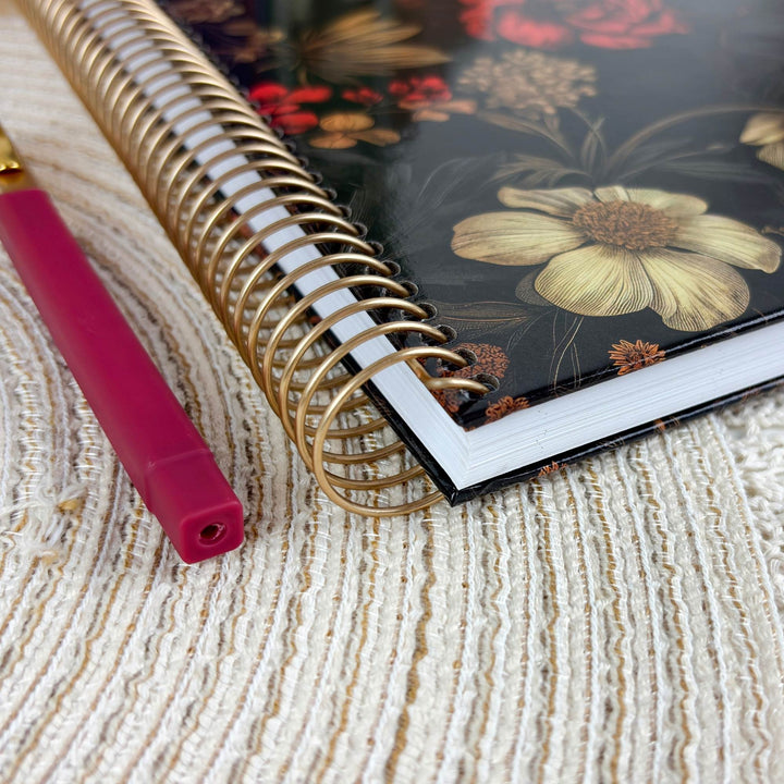A close-up of a spiral-bound notebook with a black floral cover and gold wire binding, placed on a textured cream fabric next to a pink pen.