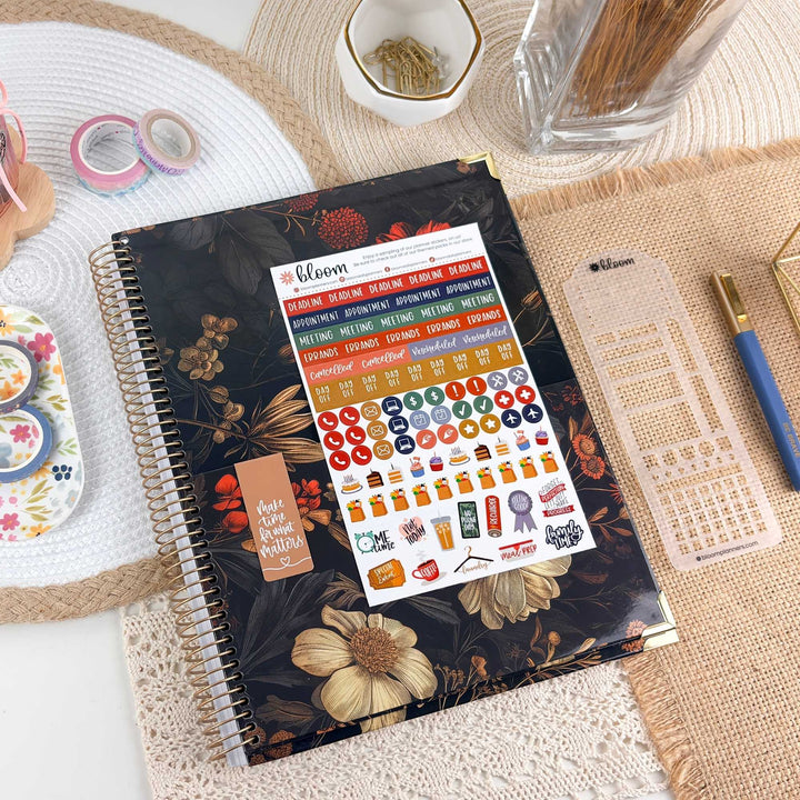 A spiral-bound planner or notebook with a dark floral cover lies on a woven mat, displaying a sheet of colorful planner stickers. The scene includes coordinating desk accessories such as washi tape, a pencil, a pen, and a small decorative item, arranged in a styled flat-lay setup.