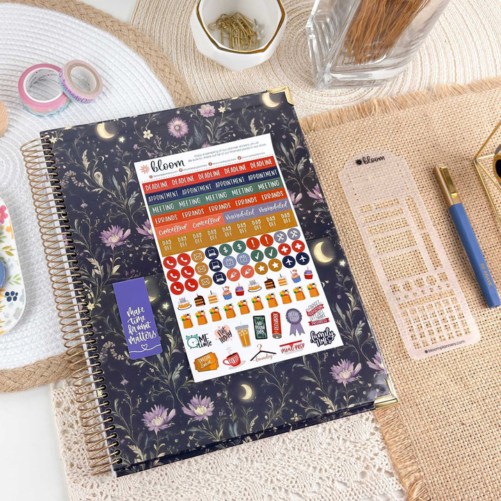 A spiral-bound planner or notebook with a dark floral cover and a sticker sheet attached, styled on a woven mat with washi tape, a pen, and other stationery around it.