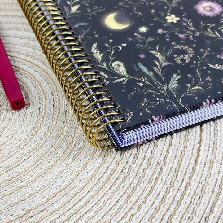 A spiral-bound notebook with a dark navy floral and celestial cover design, featuring a gold wire spiral binding, resting on a textured beige woven mat next to a pink pen.