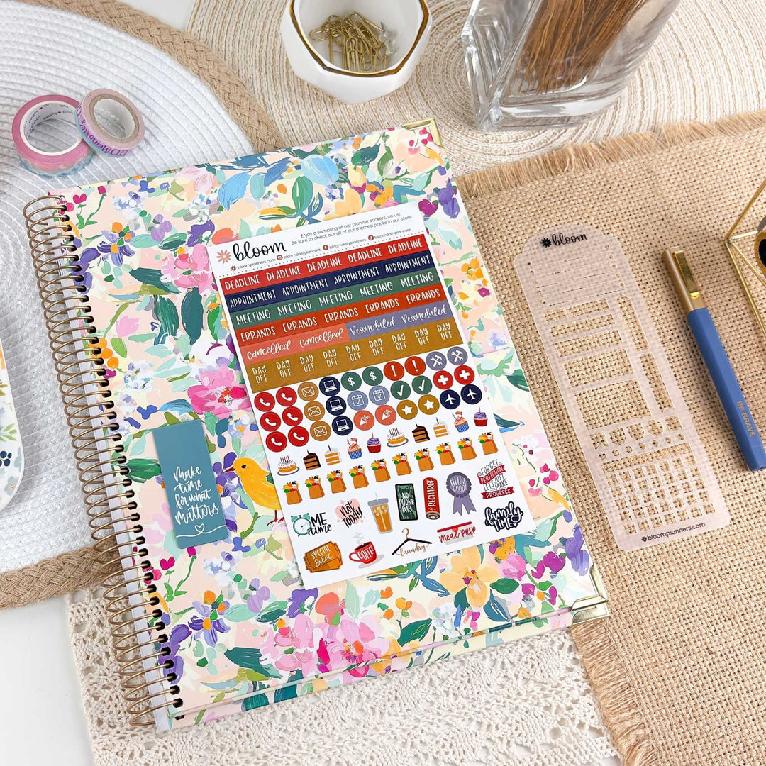 A spiral-bound planner or notebook with a colorful floral cover and a sheet of decorative stickers attached, styled on a woven mat with pens, washi tape, and other stationery around it.