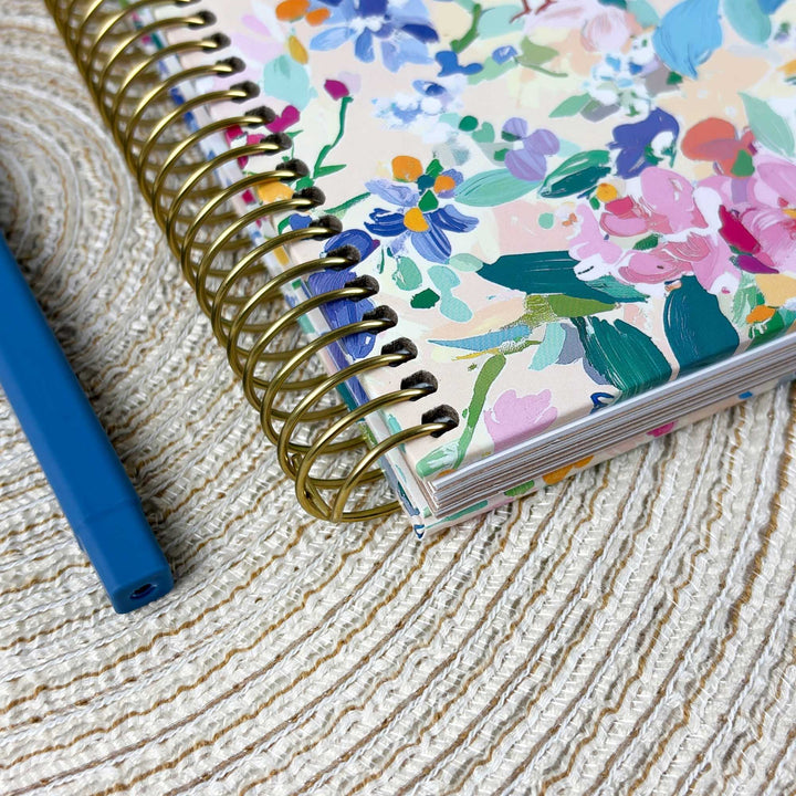 A floral spiral-bound notebook with a gold wire coil lies on a woven straw surface next to a blue pen.