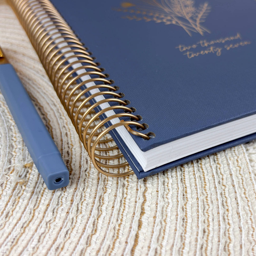 A navy blue spiral-bound notebook with gold wire binding and gold foil text, shown on a textured beige woven surface next to a matching blue pen.