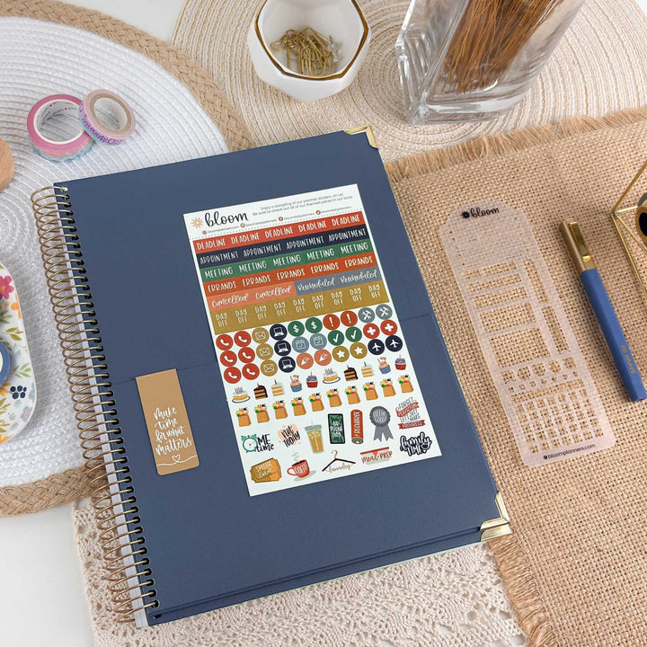 A navy blue spiral-bound planner or notebook with a decorative sticker sheet attached to the cover, styled on a woven mat with pens, tape, and other stationery around it.