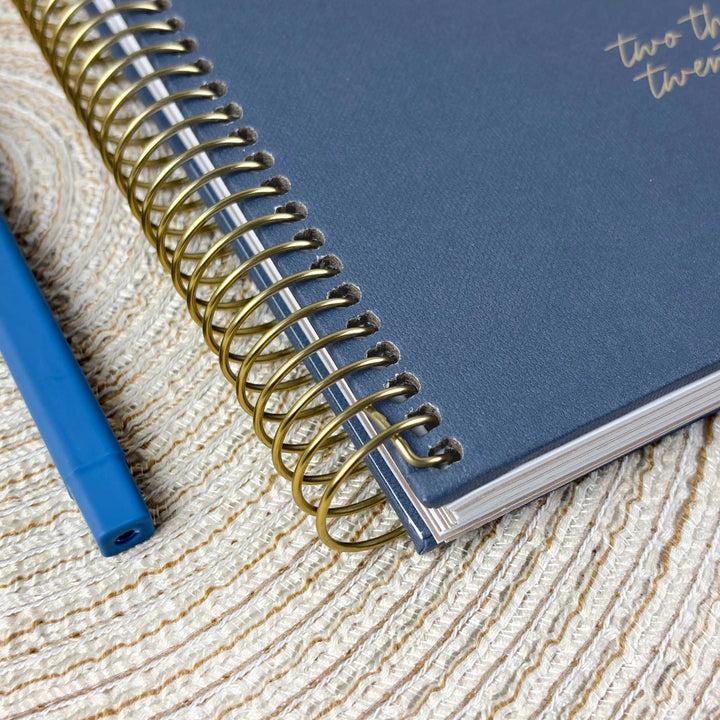 A close-up of a navy blue spiral-bound notebook with gold wire binding, placed on a textured beige woven surface next to a blue pen.