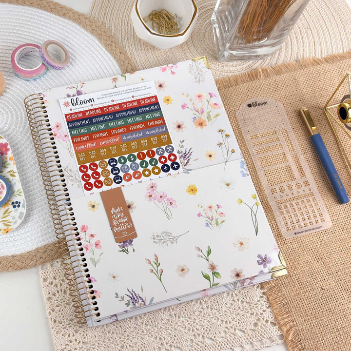 A spiral-bound planner or journal with a white floral cover lies on a woven mat, surrounded by stationery items including washi tapes, a clear plastic grid insert, a blue pen, and a small decorative item. The cover shows the brand name 'bloom' and decorative flower illustrations.